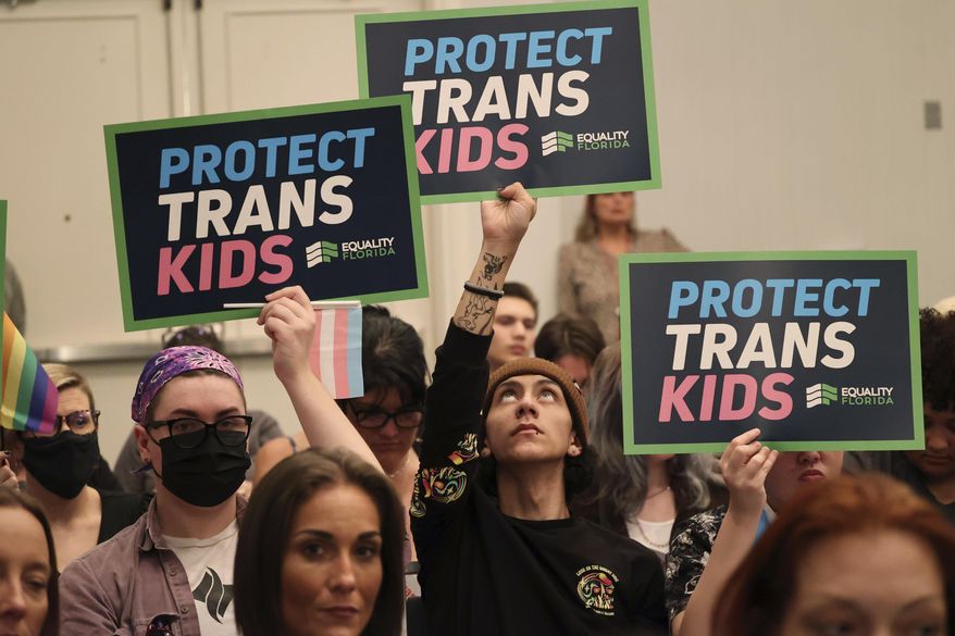 People hold signs during a joint board meeting of the Florida Board of Medicine and the Florida Board of Osteopathic Medicine, Friday, Nov. 4, 2022, in Lake Buena Vista, Fla., to establish new guidelines limiting gender-affirming care in Florida. Members have signaled support for prohibiting puberty blockers, cross-hormonal treatment and gender-affirming surgery as methods to treat gender dysphoria in patients under the age of 18. Critics say the rules, urged by Surgeon General Joseph Ladapo, would have potentially life-or-death consequences for transgender youth. (Ricardo Ramirez Buxeda/Lake Buena Vista Sentinel via AP)