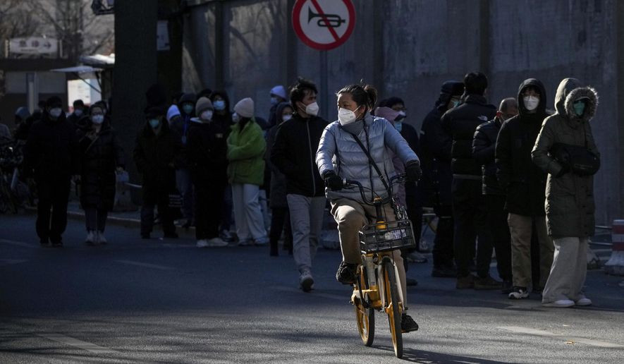 A masked woman bicycles past residents lining up to enter the fever clinic of a hospital in Beijing, Sunday, Dec. 11, 2022. Facing a surge in COVID-19 cases, China is setting up more intensive care facilities and trying to strengthen hospitals as Beijing rolls back anti-virus controls that confined millions of people to their homes, crushed economic growth and set off protests. (AP Photo/Andy Wong) **FILE**