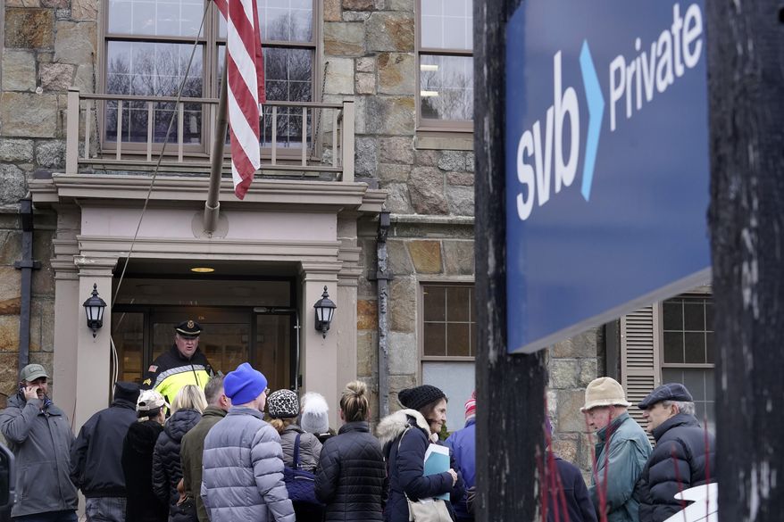 A law enforcement official, behind, stands in an entryway to a branch of Silicon Valley Bank, Monday, March 13, 2023, as customers and bystanders line up outside the branch, in Wellesley, Mass. (AP Photo/Steven Senne)
