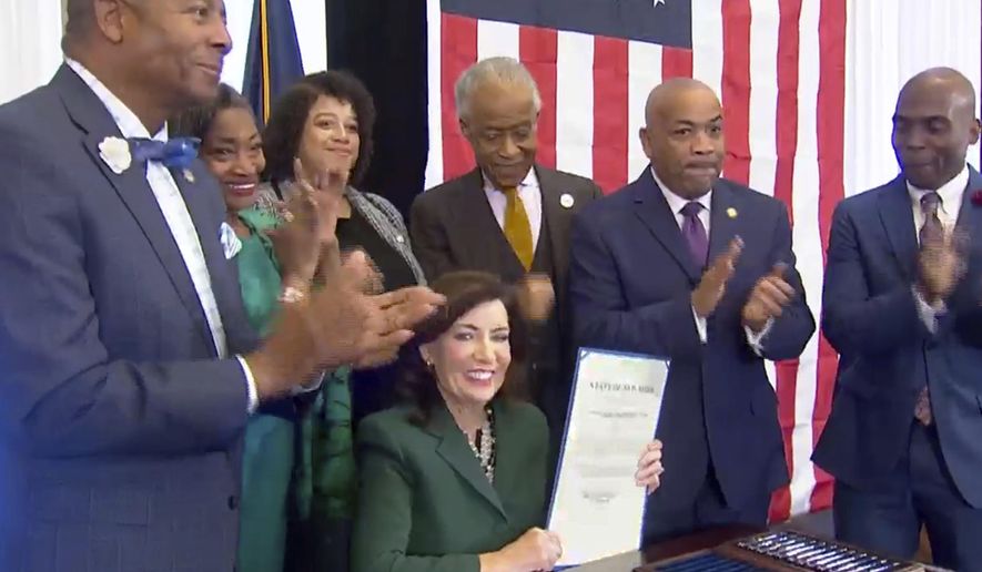 This image from video provided by the Office of The Governor shows New York Gov. Kathy Hochul, seated center, holds a bill she signed in New York, Tuesday, Dec. 19, 2023, to create a commission tasked with considering reparations to address the persistent, harmful effects of slavery in the state. She is joined by, standing from left: Dr. Yohuru Williams, Founding Director of the Racial Justice Initiative at the University of St. Thomas; Andrea Stewart-Cousins, Majority Leader of the NY State Senate; Michaelle Solages, NY State Assembly Woman; Rev. Al Sharpton; Carl Heastie, Speaker of the NY State Assembly; James Sanders, NY State Senator. (Office of the Governor via AP)