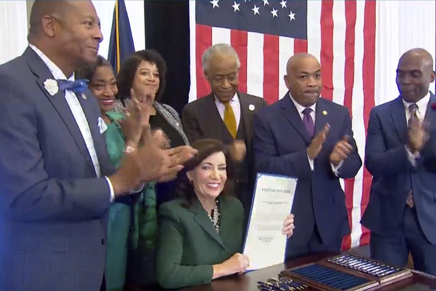 This image from video provided by the Office of The Governor shows New York Gov. Kathy Hochul, seated center, holds a bill she signed in New York, Tuesday, Dec. 19, 2023, to create a commission tasked with considering reparations to address the persistent, harmful effects of slavery in the state. She is joined by, standing from left: Dr. Yohuru Williams, Founding Director of the Racial Justice Initiative at the University of St. Thomas; Andrea Stewart-Cousins, Majority Leader of the NY State Senate; Michaelle Solages, NY State Assembly Woman; Rev. Al Sharpton; Carl Heastie, Speaker of the NY State Assembly; James Sanders, NY State Senator. (Office of the Governor via AP)