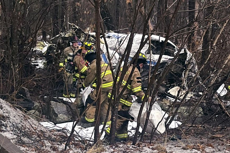 In this photo provided by Eddie Saktanaset, firefighters recover items from the site of a small plane crash, at rear, in a wooded area behind his home, Friday, Jan. 26, 2024, in Londonderry, N.H. The Wiggins Air Beechcraft 99, which was flying light cargo to Maine, crashed about 7:30 a.m. near the Manchester-Boston Regional Airport shortly after takeoff. Firefighters helped remove the trapped pilot, who was taken to a hospital, authorities said Friday. (Eddie Saktanaset photo via Associated Press)