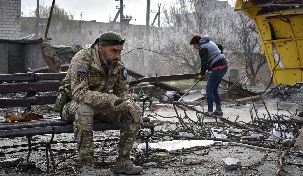 A Ukrainian serviceman smokes sitting on a bench as a local resident clears debris near a building damaged in the Russian air raid in the town of Orikhiv, Zaporizhzhia region, Ukraine, Friday, Apr. 5, 2024. (AP Photo/Andriy Andriyenko)