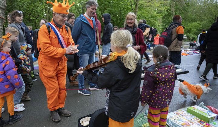 An orange-clad man applauds for a girl playing violin as his orange-clad dog, bottom right corner, runs off during King&#x27;s Day celebrations in Amsterdam, Netherlands, Saturday, April 27, 2024. (AP Photo/Peter Dejong)