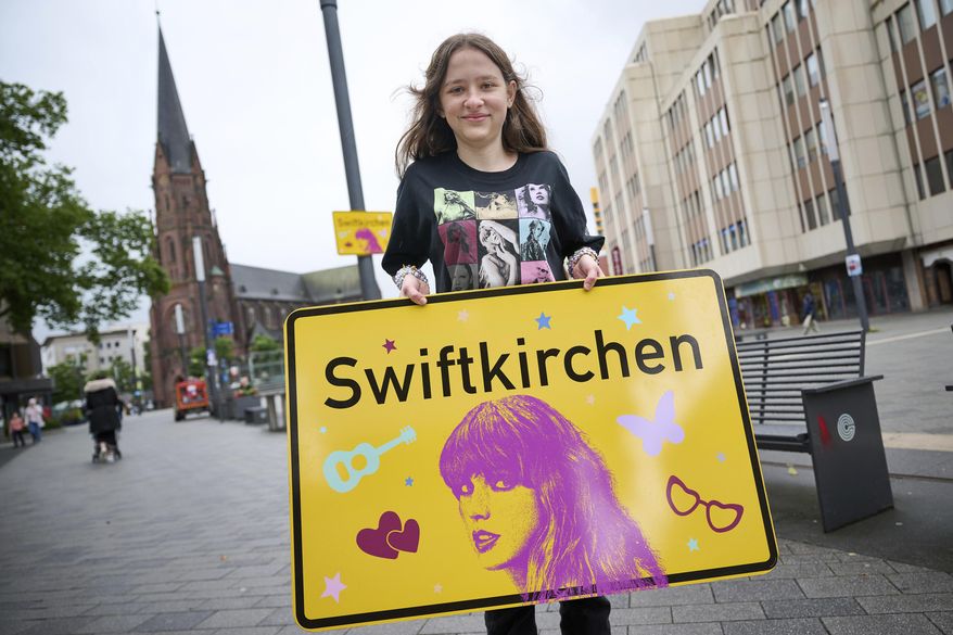 Schoolgirl Aleshanee Westhoff shows a "Swiftkirchen" town sign in honor of musician Taylor Swift in Gelsenkirchen, Germany, Tuesday, July 2, 2024. The Swifties are about to take over the German city of Gelsenkirchen, where American superstar Taylor Swift is set to give three concerts of her Eras Tour later this month. In honor of the singer, the city renamed itself “Swiftkirchen" — at least temporarily to welcome the tens of thousands of fans who are expected to come for the Eras Tour shows on July 17, 18 and 19, German news agency dpa reported. (Bernd Thissen/dpa via AP)