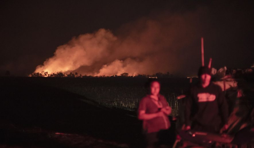 Smoke billows behind Indigenous families as part of attacks on the Guarani Kaiowa ethnicity on Panambi Lagoa Rica Indigenous land in Douradina, Mato Grosso do Sul state, Brazil, Saturday, Aug. 3, 2024. The Indigenous people allege that farmers are responsible for the attack to discourage them from recuperating land. (AP Photo/Gabriel Schlickmann)