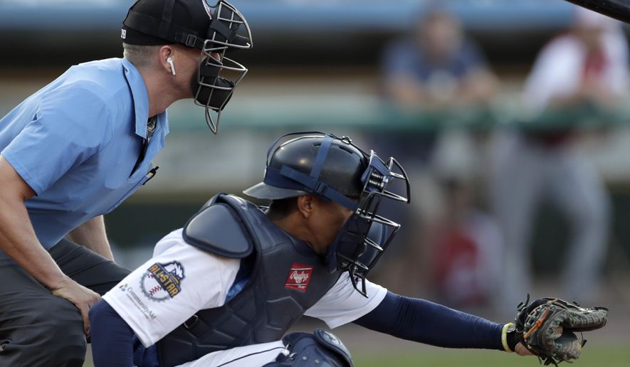 FILE -Home plate umpire Brian deBrauwere, left, huddles behind Freedom Division catcher James Skelton, of the York Revolution, as the official wears an earpiece during the first inning of the Atlantic League All-Star minor league baseball game, Wednesday, July 10, 2019, in York, Pa. Major League Baseball will test robot umpires as part of a challenge system during spring training at 13 ballparks hosting 19 teams, which could lead to regular-season use in 2026. (AP Photo/Julio Cortez, File)