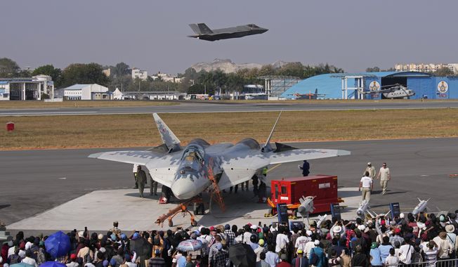 Visitors watch as U.S. Air Force fighter aircraft F-35 flies over Russia's Su-57 fighter aircraft, parked at the static display area, on the fourth day of the Aero India 2025, a biennial event, at Yelahanka air base in Bengaluru, India, Thursday, Feb. 13, 2025. (AP Photo/Aijaz Rahi)