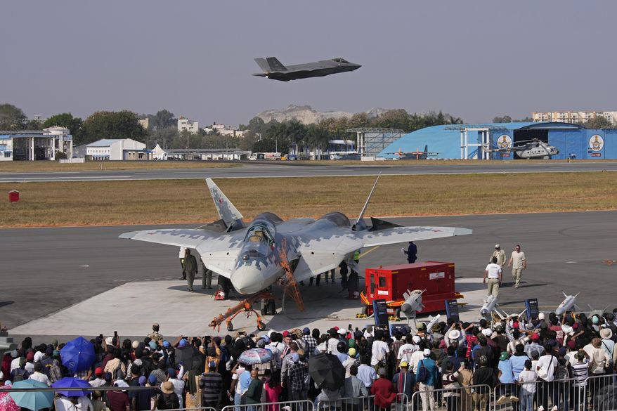 Visitors watch as U.S. Air Force fighter aircraft F-35 flies over Russia's Su-57 fighter aircraft, parked at the static display area, on the fourth day of the Aero India 2025, a biennial event, at Yelahanka air base in Bengaluru, India, Thursday, Feb. 13, 2025. (AP Photo/Aijaz Rahi)