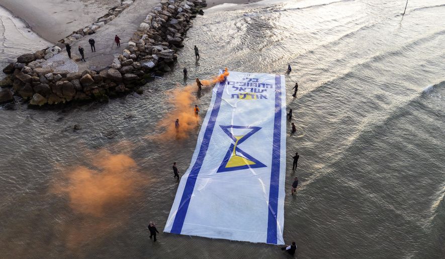 Relatives and supporters of Israelis held hostage by Hamas in Gaza mark 500 days of their captivity by spreading a massive Israeli flag depicting an hourglass in the Mediterranean Sea, in Tel Aviv, Israel, Monday, Feb. 17, 2025. Hebrew of the flag reads "Without the abductees, Israel runs out". (AP Photo/Oded Balilty)