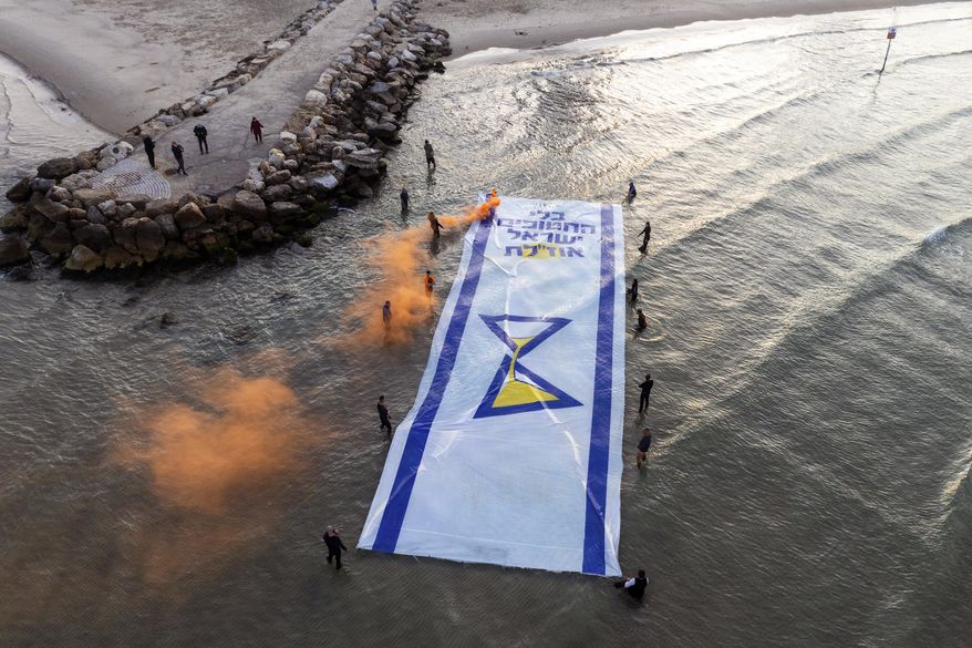 Relatives and supporters of Israelis held hostage by Hamas in Gaza mark 500 days of their captivity by spreading a massive Israeli flag depicting an hourglass in the Mediterranean Sea, in Tel Aviv, Israel, Monday, Feb. 17, 2025. Hebrew of the flag reads "Without the abductees, Israel runs out". (AP Photo/Oded Balilty)
