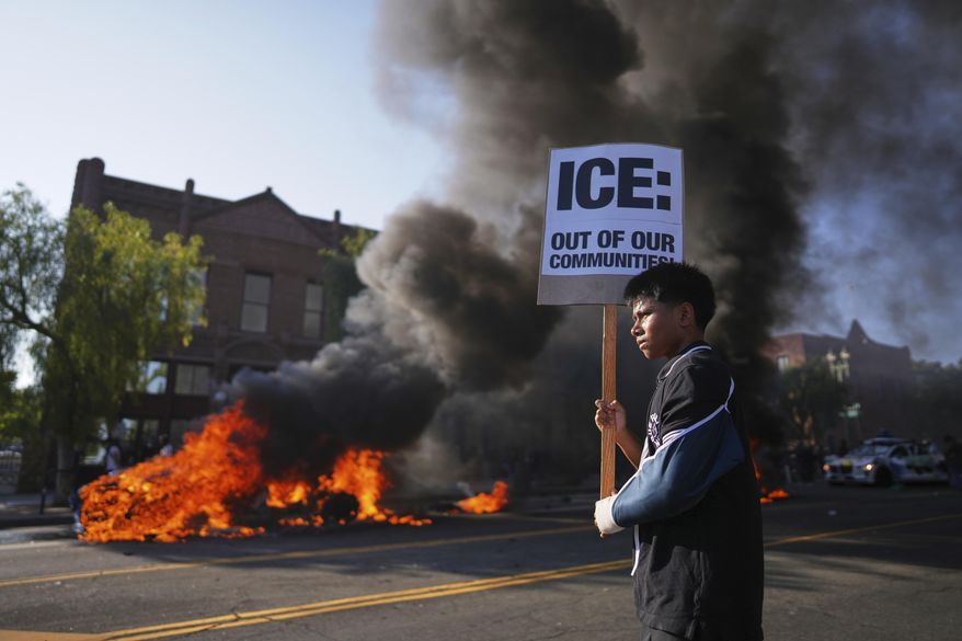 A protester holds a sign as a Waymo taxi burns near the metropolitan detention center of downtown Los Angeles, Sunday, June 8, 2025, following last night's immigration raid protest. (AP Photo/Eric Thayer)