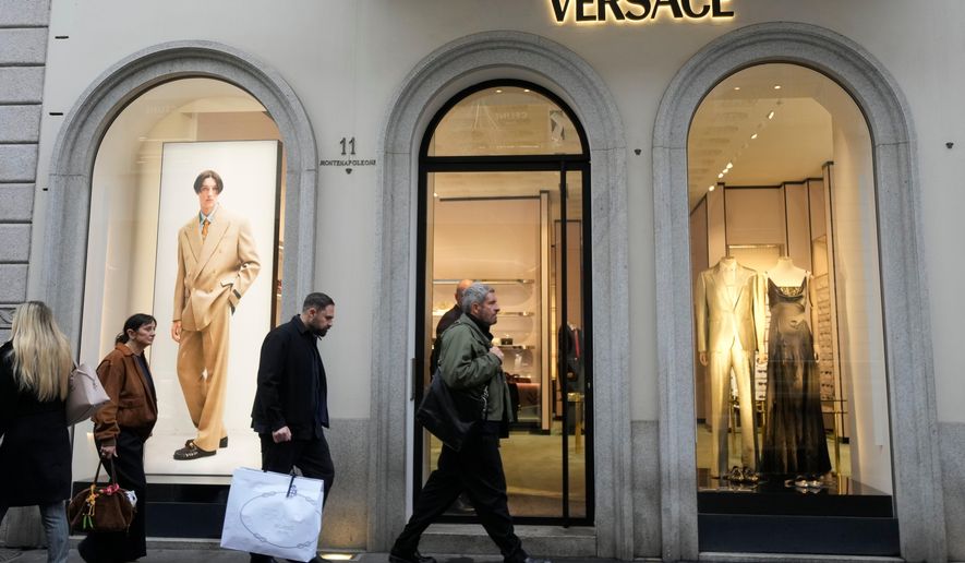 A man walks with a Prada shopping bag in front of a Versace shop, at the Montenapoleone luxury fashion street, in Milan, Italy, Tuesday, Dec. 2, 2025. (AP Photo/Luca Bruno)