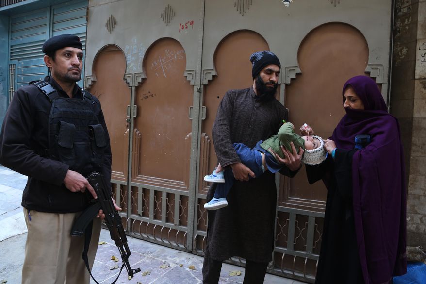 A police officer stands guard as a health worker, right, administers a polio vaccine to a child at a neighbourhood in Peshawar, Pakistan, Monday, Monday, Dec. 15, 2025. (AP Photo/Muhammad Sajjad)