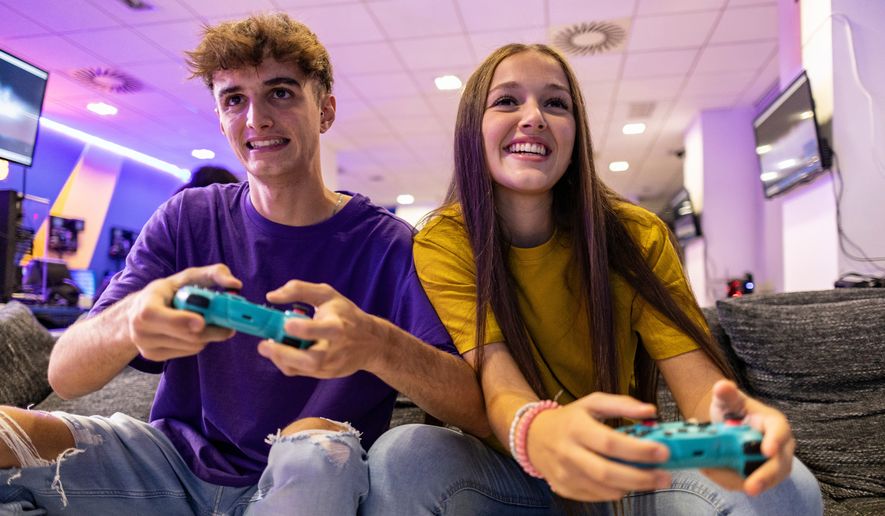 Two young gamers sit on a couch in a gaming room, holding wireless controllers and playing video games. File photo credit: PintoArt via Shutterstock.