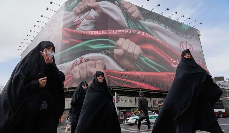 Women, one flashing a victory hand gesture, cross a street under a huge banner showing hands firmly holding Iranian national flags as a sign of patriotism, in Tehran, Iran, Jan. 14, 2026. (AP Photo/Vahid Salemi, File)