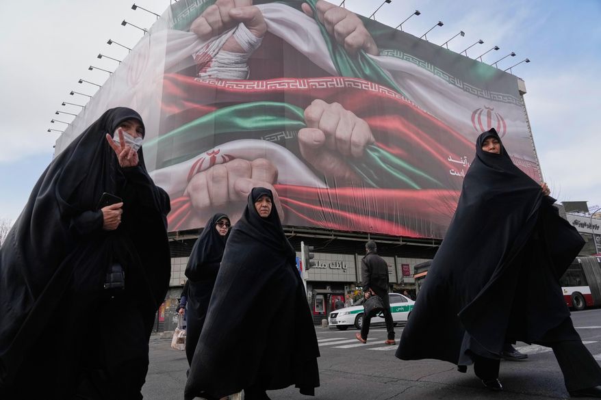 Women, one flashing a victory hand gesture, cross a street under a huge banner showing hands firmly holding Iranian national flags as a sign of patriotism, in Tehran, Iran, Jan. 14, 2026. (AP Photo/Vahid Salemi, File)