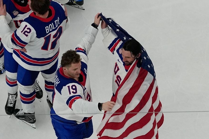United States' Matthew Tkachuk (19) helps put an American flag around goalie Connor Hellebuyck after the United States beat Canada in overtime to win the men's gold medal hockey game at the 2026 Winter Olympics, in Milan, Italy, Sunday, Feb. 22, 2026. (AP Photo/David J. Phillip)