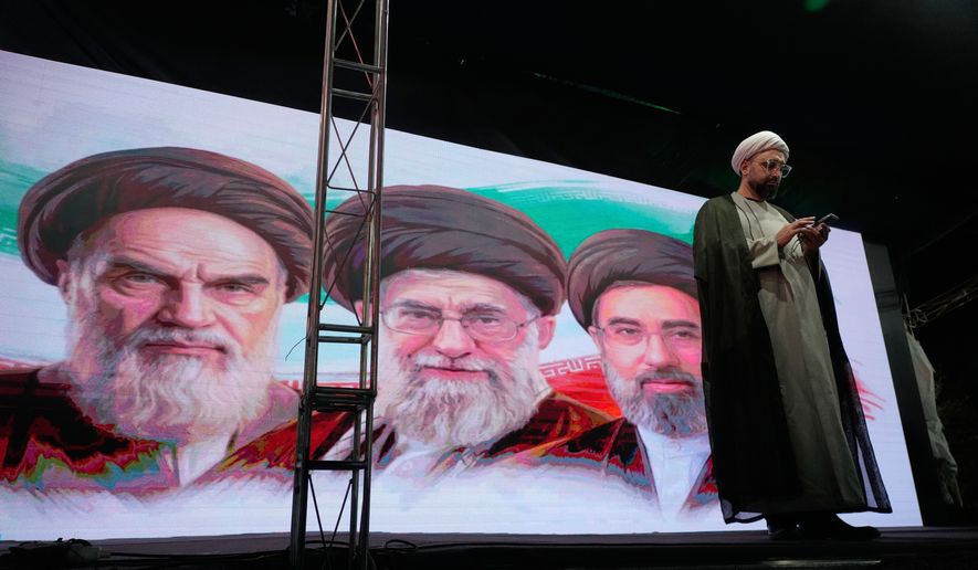 A cleric checks his cell phone on stage in front of a screen displaying portraits of the late Iranian revolutionary founder Ayatollah Khomeini, left, late Supreme Leader Ayatollah Ali Khamenei, center, and Supreme Leader Ayatollah Mojtaba Khamenei, during a pro-government gathering in Tehran, Iran, Sunday, June 7, 2026. (AP Photo/Vahid Salemi)