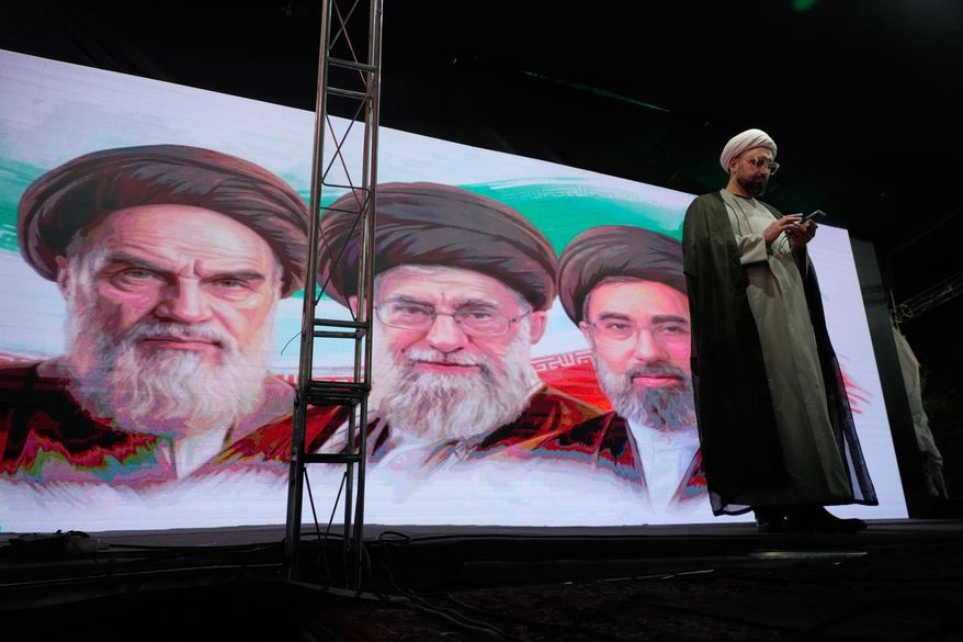 A cleric checks his cell phone on stage in front of a screen displaying portraits of the late Iranian revolutionary founder Ayatollah Khomeini, left, late Supreme Leader Ayatollah Ali Khamenei, center, and Supreme Leader Ayatollah Mojtaba Khamenei, during a pro-government gathering in Tehran, Iran, Sunday, June 7, 2026. (AP Photo/Vahid Salemi)