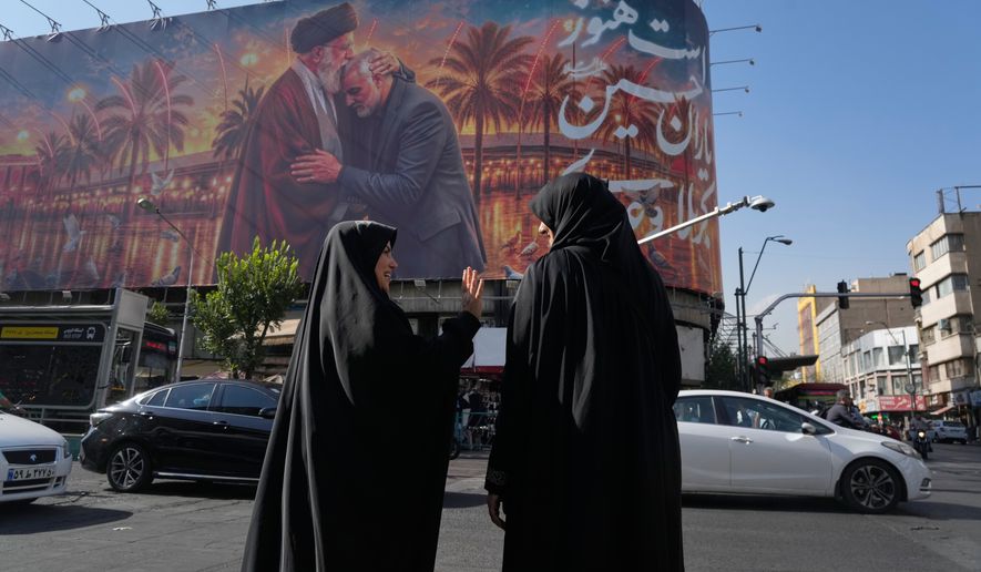 Women talk in front of a banner with graphic depicting the late Iranian Supreme Leader Ayatollah Ali Khamenei kissing head of the late commander of Iran's Revolutionary Guard expeditionary Quds Force, Gen. Qassem Soleimani, who was killed in a U.S. drone attack in 2020 in Iraq, as they wait for the green light to cross an intersection in downtown Tehran, Iran, Monday, June 29, 2026. (AP Photo/Vahid Salemi)