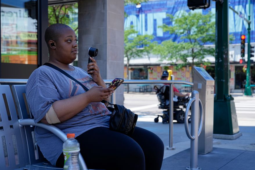 Simone Mosley waits for their rideshare in shade at a bus stop during an extreme heat warning Monday, July 13, 2026, in Minneapolis. (AP Photo/Ellen Schmidt)