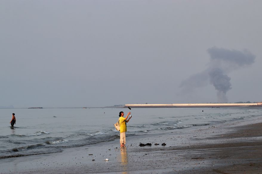 A woman stands at the water's edge along the Strait of Hormuz as a plume of smoke rises in the background following an explosion, off Bandar Abbas, Iran, Monday, July 13, 2026. (Razieh Poudat/ISNA via AP)