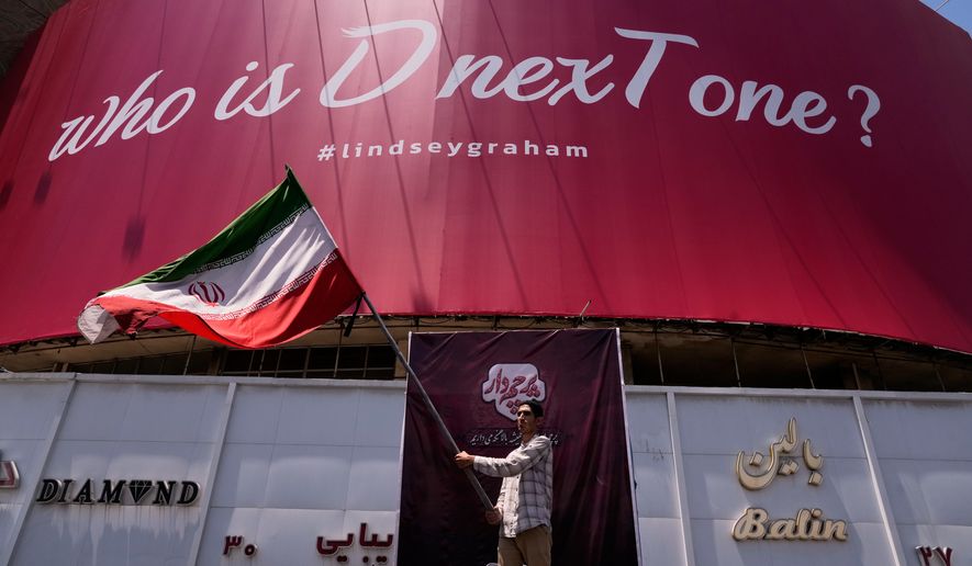 A man waves an Iranian flag beneath a billboard reading in English, "Who is D nexT one?" and "#lindseygraham," referring to late U.S. Sen. Lindsey Graham and using the capital letters "D" and "T" in an apparent play on the initials of U.S. President Donald Trump, in downtown Tehran, Iran, Thursday, July 16, 2026. (AP Photo/Vahid Salemi)
