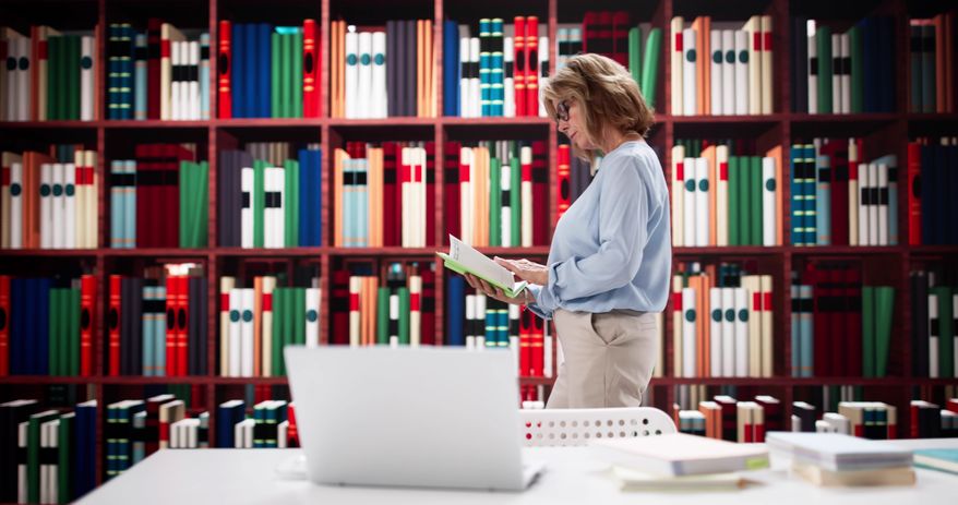 Female professor researching In university library with a laptop and a book. File photo credit: Andrey_Popov via Shutterstock.