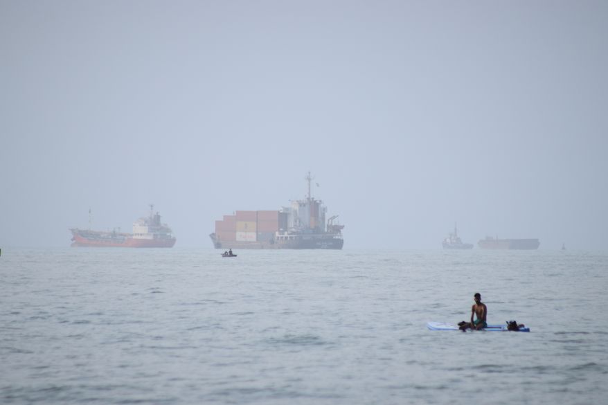 A man sits on a surfboard as a container ship and other commercial vessels appear anchored in the Strait of Hormuz off Bandar Abbas, Iran, Monday, July 27, 2026. (Razieh Poudat/ISNA via AP, File)