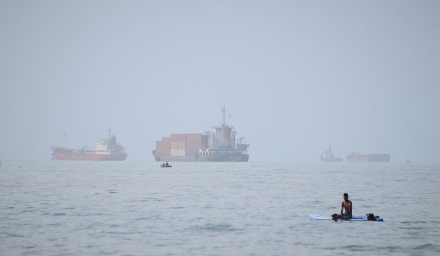 A man sits on a surfboard as a container ship and other commercial vessels appear anchored in the Strait of Hormuz off Bandar Abbas, Iran, Monday, July 27, 2026. (Razieh Poudat/ISNA via AP)