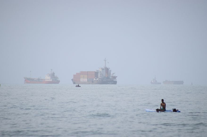A man sits on a surfboard as a container ship and other commercial vessels appear anchored in the Strait of Hormuz off Bandar Abbas, Iran, Monday, July 27, 2026. (Razieh Poudat/ISNA via AP)