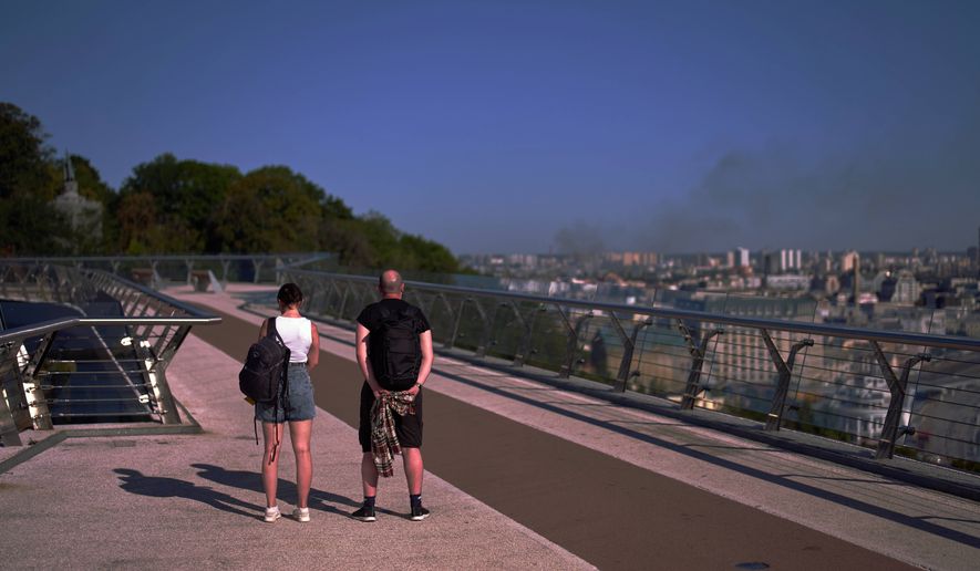 People observe a daily nationwide minute of silence in memory of soldiers killed in the war with Russia as smoke rises from the city following a Russian missile attack in Kyiv, Sunday, Aug. 16, 2026. (AP Photo/Francisco Seco)