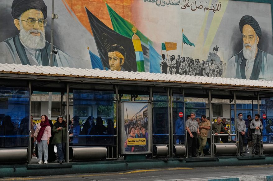 Commuters stand at a bus stop on Islamic Revolution street in front of a mural depicting the late Iranian revolutionary founder Ayatollah Khomeini, right, the late Supreme Leader Ayatollah Ali Khamenei, who was killed in the U.S. and Israel strike on Feb. 28, left, and Basij paramilitary forces, in Tehran, Iran, Tuesday, Oct. 6, 2026. (AP Photo/Vahid Salemi)