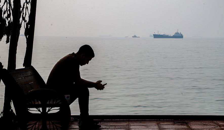 A man checks his cellphone at a waterfront shelter as commercial vessels appear anchored in the Strait of Hormuz off Bandar Abbas, Iran, Monday, Tuesday, Oct, 6, 2026. (Amirhosein Khorgooi/ISNA via AP)