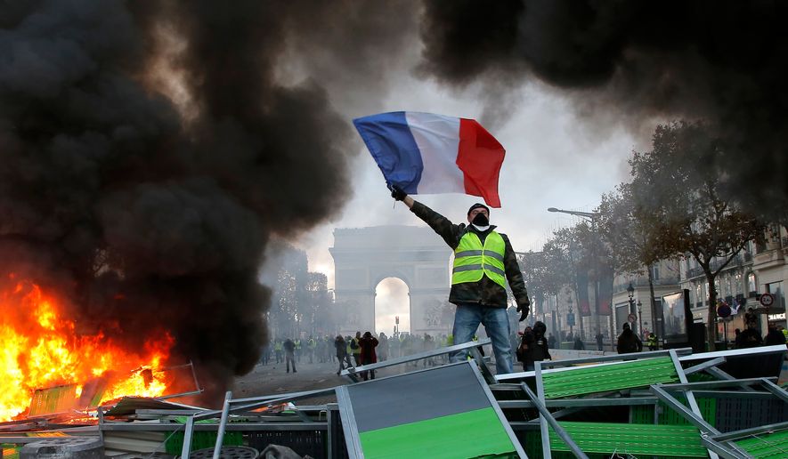 FILE - A demonstrator wearing a yellow vest waves the French flag on top of a burning barricade on the Champs-Elysees avenue with the Arc de Triomphe in background in Paris, on Saturday, Nov. 24, 2018, during a demonstration against the rising fuel taxes. (AP Photo/Michel Euler, File)