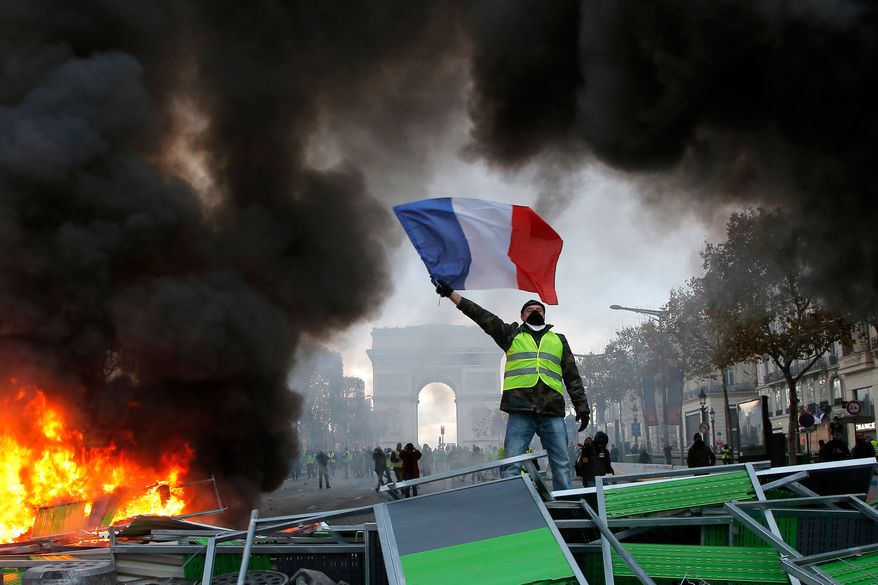 FILE - A demonstrator wearing a yellow vest waves the French flag on top of a burning barricade on the Champs-Elysees avenue with the Arc de Triomphe in background in Paris, on Saturday, Nov. 24, 2018, during a demonstration against the rising fuel taxes. (AP Photo/Michel Euler, File)