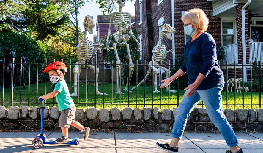 Sam McNamara, 3, of Washington, left, followed by his grandmother, rides by a yard decorated with giant skeletons, both human and dinosaur, Oct. 14, 2020, in advance celebration of Halloween in Washington. (AP Photo/Jacquelyn Martin, File)