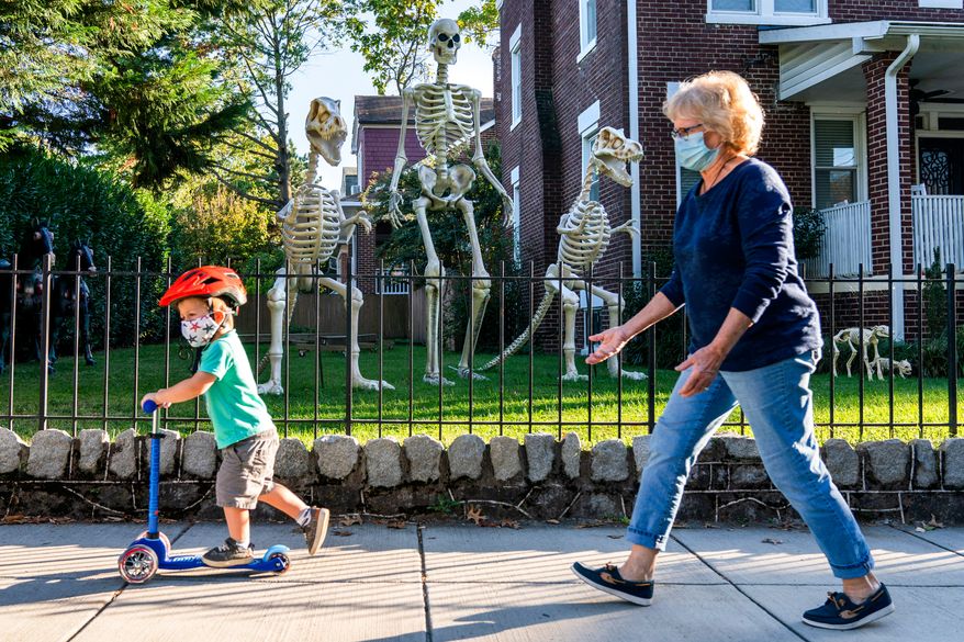 Sam McNamara, 3, of Washington, left, followed by his grandmother, rides by a yard decorated with giant skeletons, both human and dinosaur, Oct. 14, 2020, in advance celebration of Halloween in Washington. (AP Photo/Jacquelyn Martin, File)