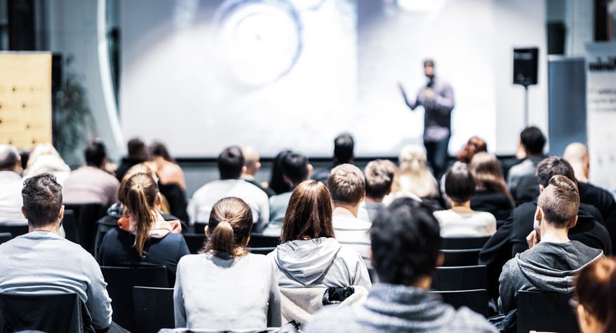 Speaker giving a talk in a conference hall. File photo credit: Matej Kastelic via Shutterstock.